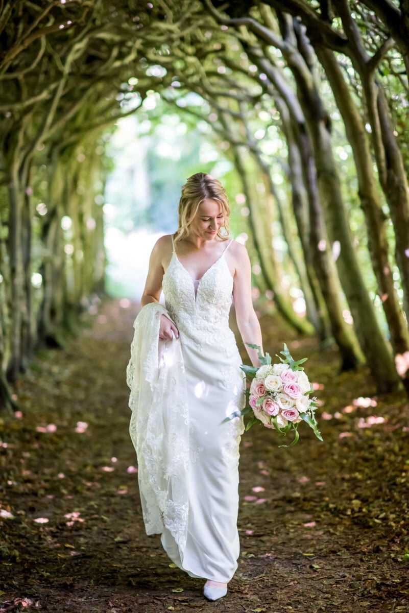 Bruiloftsfoto van bruid in witte kanten trouwjurk met roze en wit boeket, lopend onder groene bomen in zonlicht; romantische sfeer, trouwreportage buitenlocatie.