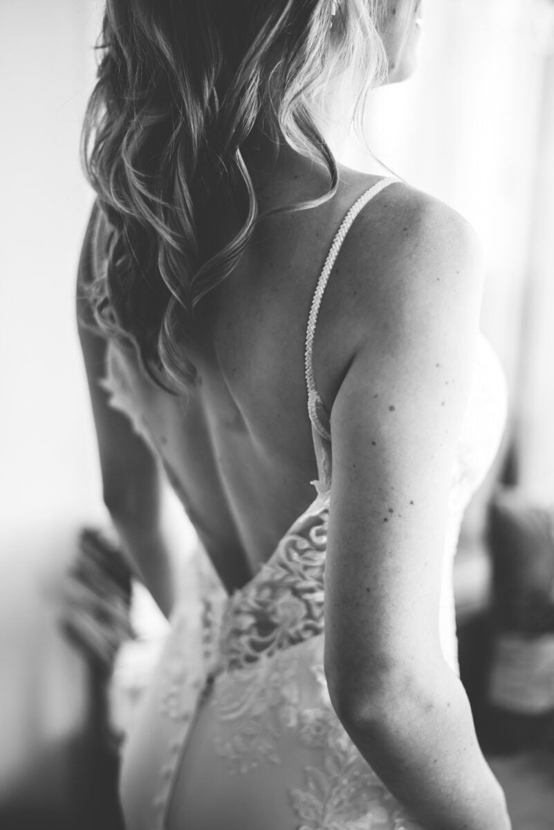 Zwart-wit foto van een vrouw in kanten trouwjurk met open rug en dunne bandjes, golvend haar over schouder, natuurlijk licht synthetische jurktextuur en huid – bruidsmode inspiratie.