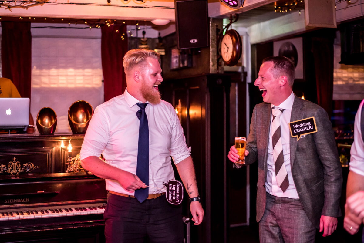 Twee mannen in nette kleding lachen samen op een bruisend feest of bruiloft, met bier, verkleedaccessoires, piano, DJ en sfeervolle lichtjes op de achtergrond.
