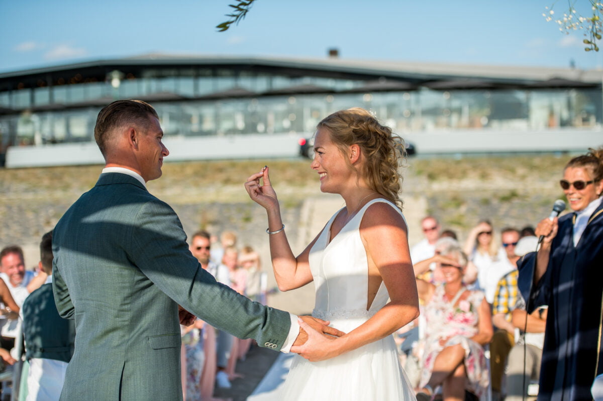 Een bruid en bruidegom houden elkaars hand vast en glimlachen naar elkaar tijdens een huwelijksceremonie in de open lucht, met gasten op de achtergrond en een ambtenaar die in een microfoon spreekt.