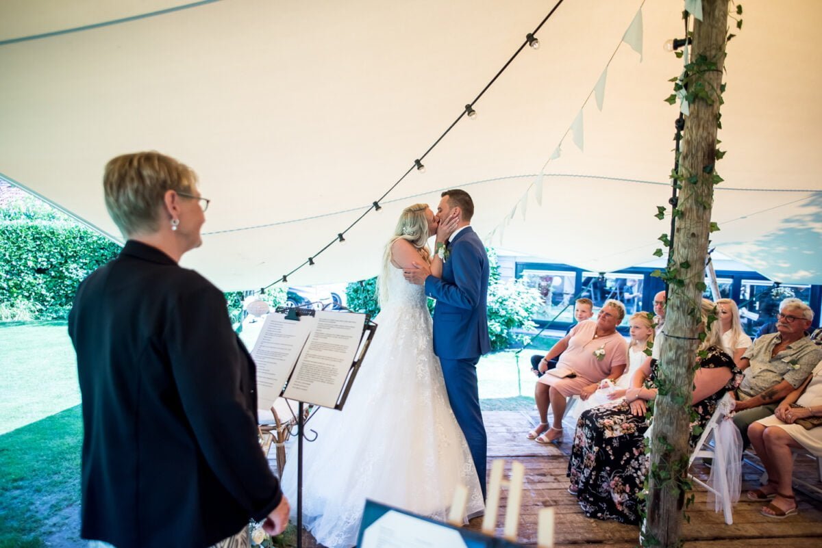 Een bruid en bruidegom kussen elkaar onder een tent versierd met lichtslingers, getuige een officiant en gasten tijdens een openluchtbruiloft. De gasten zitten in rijen en het gebied is omgeven door weelderig groen.