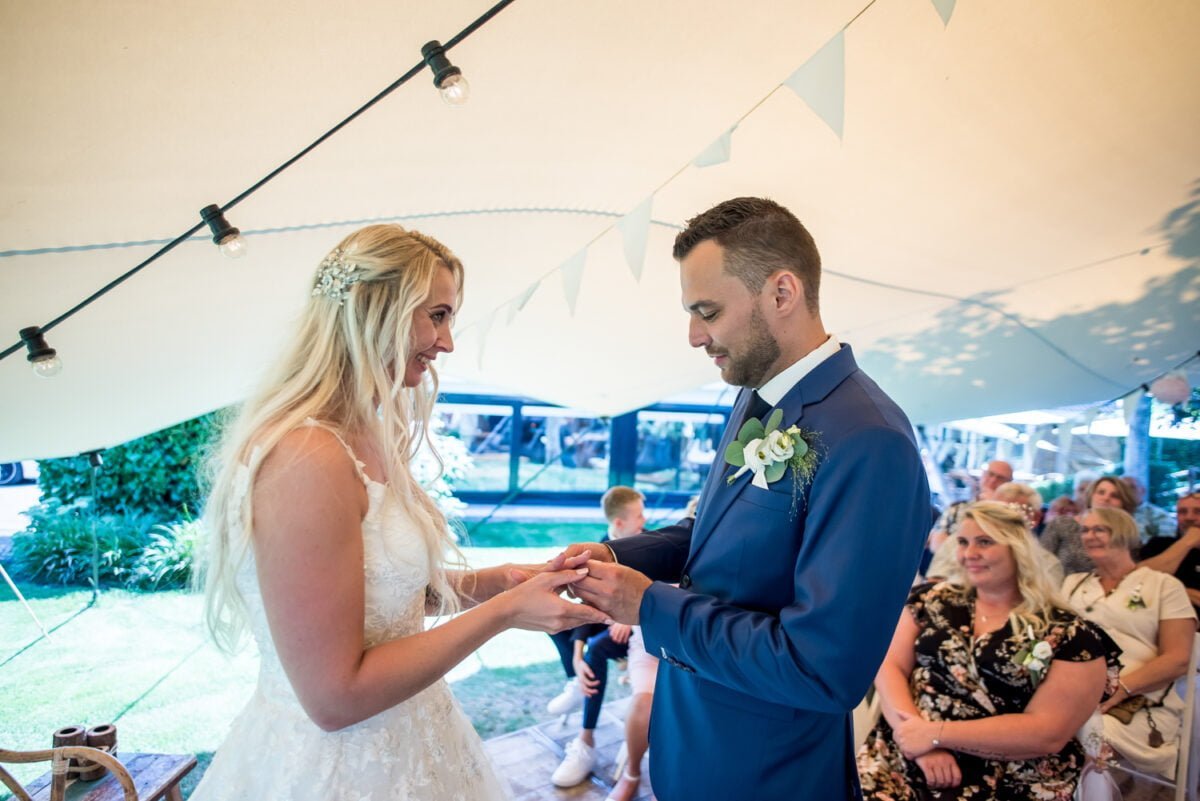 Een bruid en bruidegom wisselen tijdens hun bruiloft ringen uit onder een tent versierd met lichtslingers en witte slingers. De bruid, in een witte jurk en een gebloemd hoofddeksel, glimlacht naar de bruidegom, die gekleed is in een marineblauw pak met een bloemencorssette. Gasten kijken naar het vreugdevolle moment