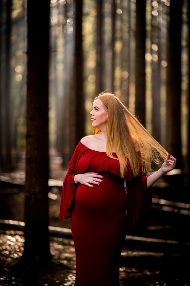 Een zwangere vrouw in een golvende rode jurk staat gracieus in een zonovergoten bos, houdt zachtjes haar buik vast en kijkt naar de zijkant, terwijl haar lange blonde haar over haar rug valt terwijl ze zich voorbereidt op haar aanstaande bruiloft.