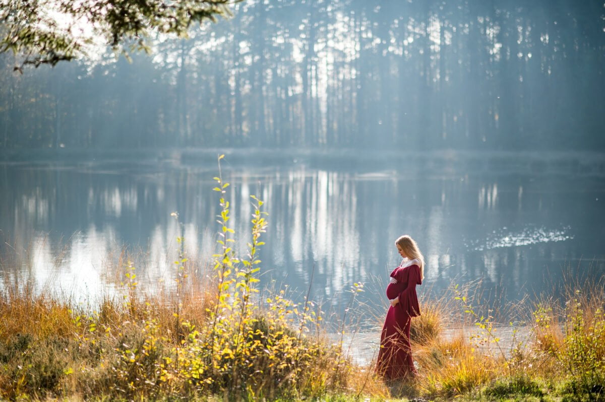 Een vrouw in een rode jurk staat naast een rustig meer, omringd door pijnbomen en badend in zacht ochtendzonlicht, reflecterend op het rustige wateroppervlak en legt het perfecte moment vast voor haar bruiloftsfotoshoot.