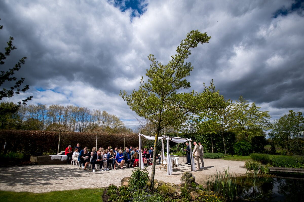 Buiten vindt een huwelijksceremonie plaats onder een dramatische, bewolkte hemel. Gasten zitten tegenover een altaar versierd met witte gordijnen. Het echtpaar staat naast de officiant, omgeven door weelderig groen en een kleine vijver die het tafereel weerspiegelt. Bomen en struiken omlijsten de schilderachtige omgeving.