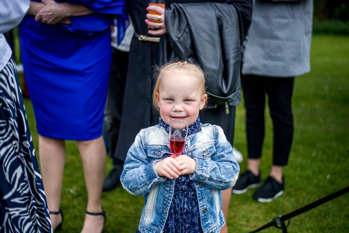 Een jong meisje met blond haar, gekleed in een spijkerjasje, glimlacht terwijl ze een glas rood sap vasthoudt. Ze staat op een grasveld omringd door een groep volwassenen, sommigen gekleed in formele kleding met wazige gezichten. De scène lijkt een bijeenkomst of evenement in de buitenlucht te zijn.