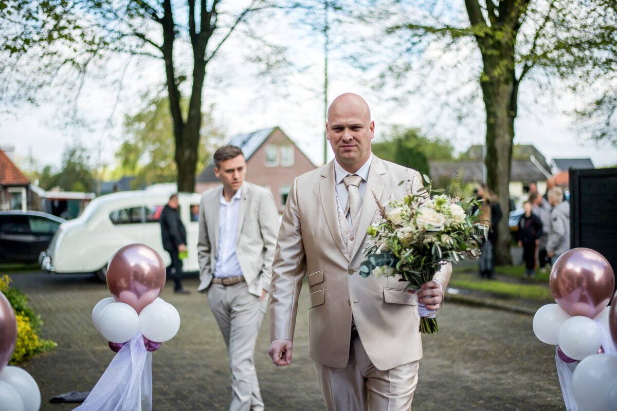 Een man in een lichtbeige pak en stropdas, met een boeket witte en groene bloemen in zijn hand, loopt over een pad versierd met ballonnen. Een andere man in een soortgelijk pak volgt hem. Bomen en een oldtimer staan op de achtergrond, wat een buitenhuwelijkssetting suggereert.