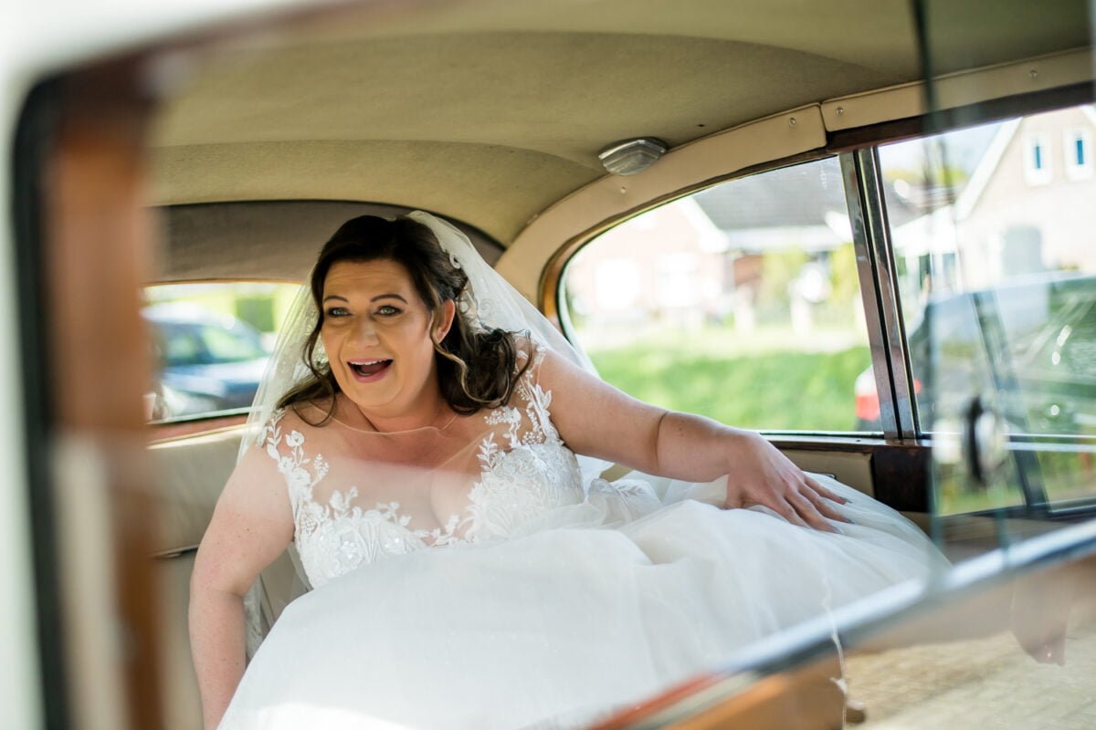 Een bruid met donker, golvend haar en een witte kanten bruidsjurk zit in een vintage auto. Ze glimlacht en kijkt naar het raam, haar sluier zachtjes over haar schouders gedrapeerd en haar hand rust op de stoel. Huizen en groen zijn zichtbaar door het autoraam op de achtergrond.