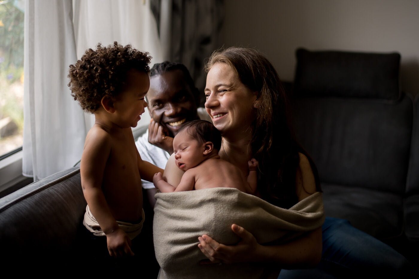 Een vrolijk familietafereel met een vrouw en een man die op een bank zitten en naar elkaar glimlachen. De vrouw wiegt een slapende pasgeborene, gewikkeld in een deken. Een peuter met krullend haar staat vlakbij, zonder shirt, en maakt contact met de volwassenen. Zacht licht uit een raam versterkt de warme sfeer.