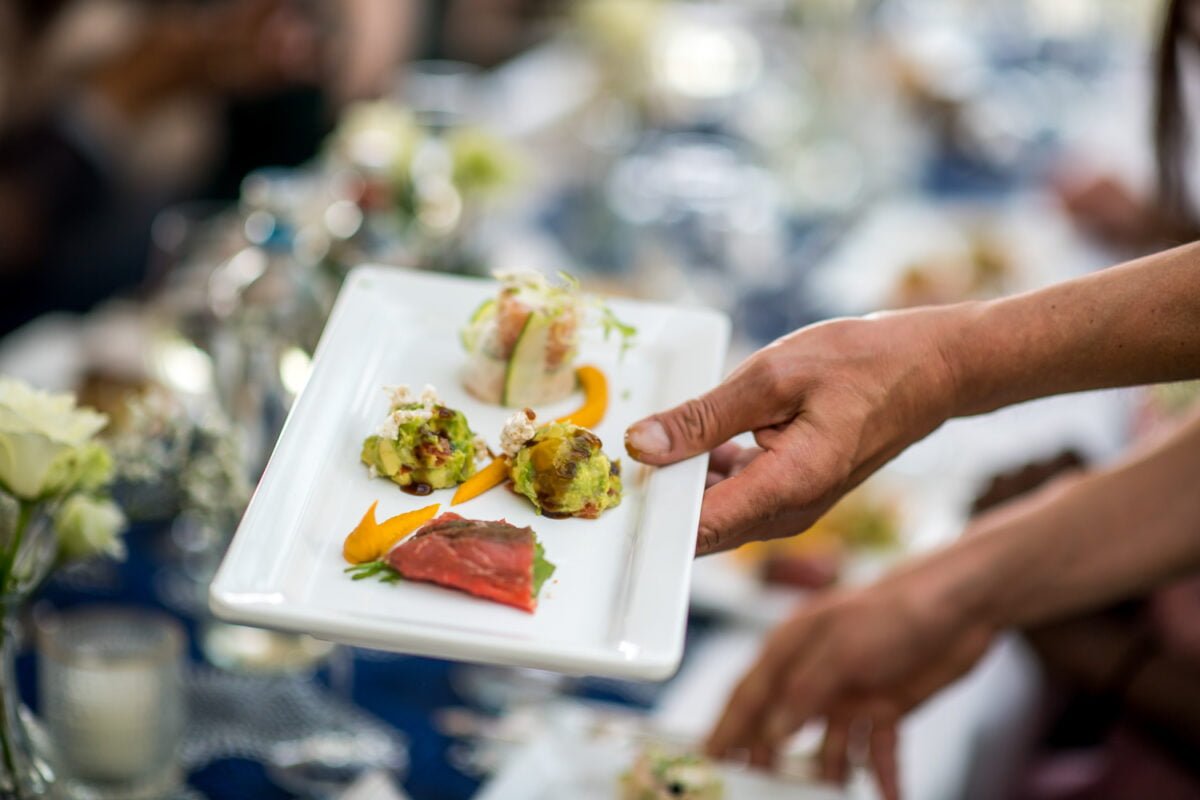 Een hand houdt een rechthoekig wit bord vast met een assortiment elegant opgemaakte hapjes. Het gerecht bevat kleine porties van verschillende gerechten, waaronder een stuk vlees met rode saus, een groene groentemix en nog twee andere kunstig gerangschikte items. Een eettafel is wazig op de achtergrond.