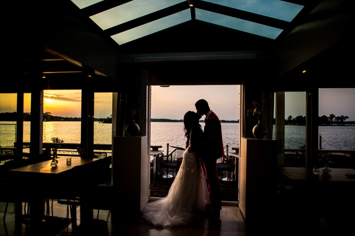 De afgetekende bruid en bruidegom delen een teder moment, omlijst door een zonovergoten deuropening die leidt naar een patio aan het meer. Binnen creëren slecht verlichte tafels een intieme sfeer, terwijl de gouden zonsondergang het serene water op de achtergrond verlicht en een romantische gloed over het tafereel werpt.