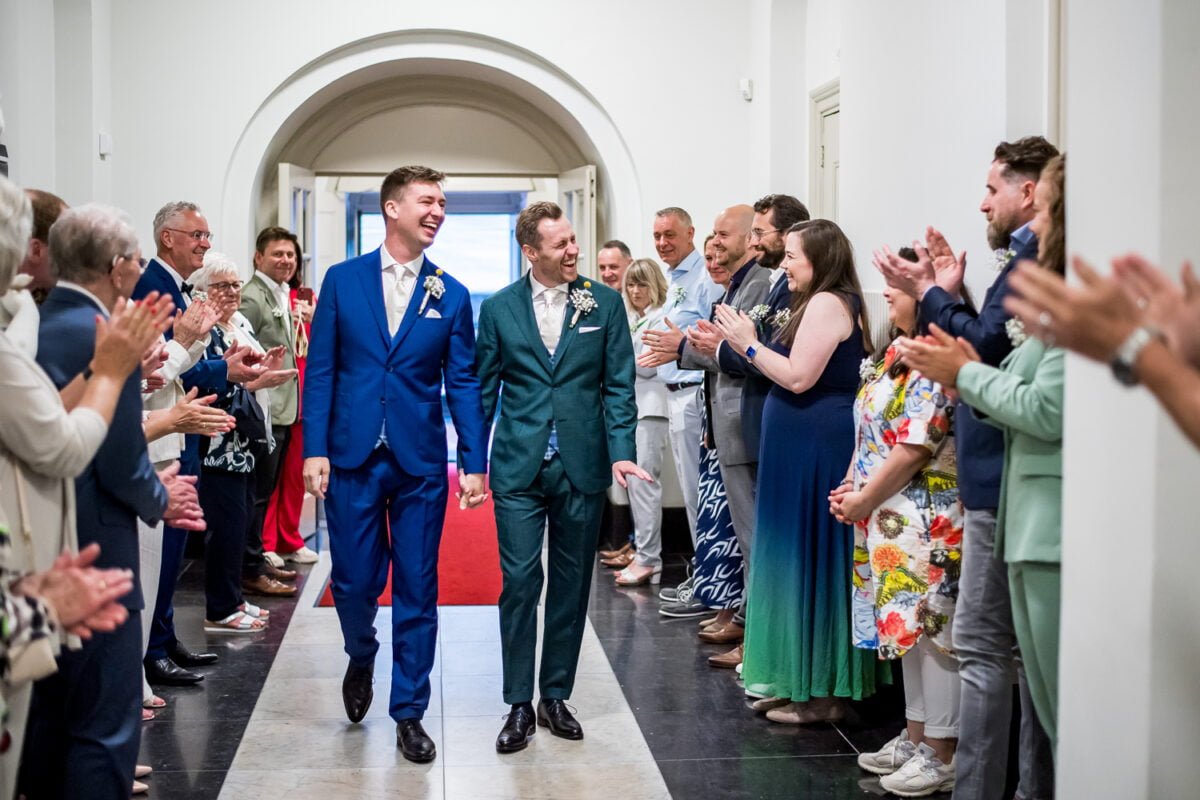 Twee bruidegommen, de een in een blauw pak en de ander in een groen pak, lopen hand in hand door een gang vol applaudisserende gasten. Beide bruidegommen glimlachen vreugdevol. De gang is helder verlicht en heeft witte muren en gebogen deuropeningen, met aan het uiteinde een rode loper zichtbaar.