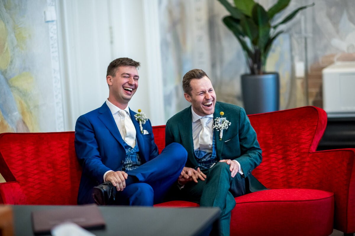 Twee mannen in pak, de een in het blauw en de ander in het groen, zitten op een rode bank en lachen hartelijk. Beiden hebben boutonnières met witte bloemen op hun jasjes gespeld. Hun lichaamstaal en uitdrukkingen suggereren vreugde en kameraadschap in een comfortabele, elegante omgeving.