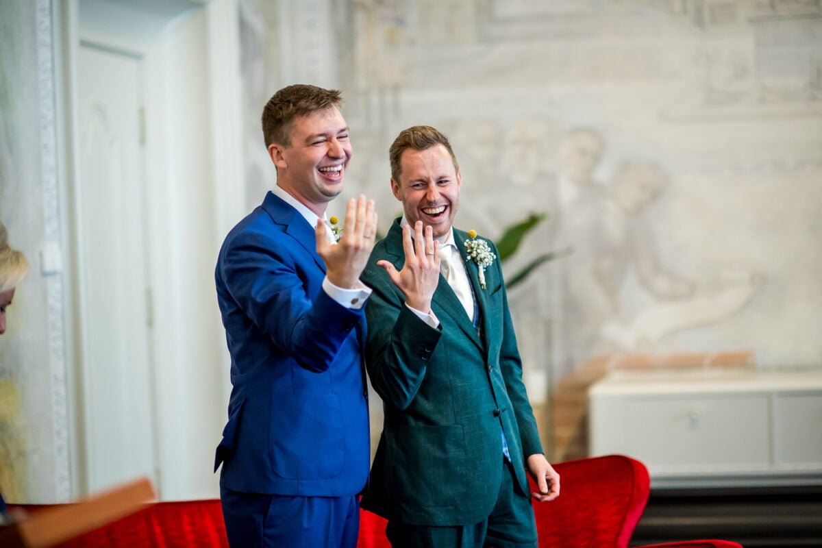 Twee mannen, allebei glimlachend en gekleed in pak, tonen hun linkerhand met trouwringen, waarschijnlijk om hun huwelijk te vieren. De een draagt een blauw pak en de ander een groen pak, elk versierd met boutonnières. Ze staan in een kamer met lichtgekleurde muren en een muurschildering op de achtergrond.