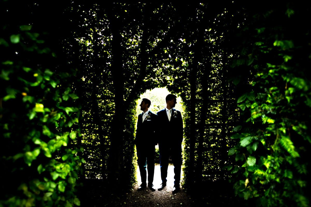 Twee mannen in pak staan zij aan zij onder een boog gevormd door weelderig groen gebladerte. De verlichting werpt ze in silhouet en benadrukt het levendige groen om hen heen. Ze staan op een pad dat in de schaduw verdwijnt, waardoor een dramatisch contrast ontstaat tussen licht en donker.