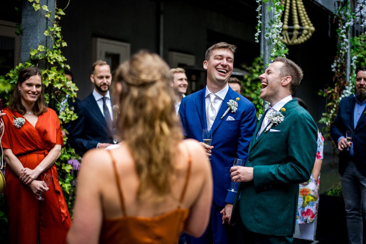 Een vrolijke huwelijksscène met twee bruidegommen in blauwe en groene pakken, die naast elkaar staan, lachen en champagneglazen vasthouden. Gasten omringen hen glimlachend; een vrouw in een oranje jurk staat met haar rug naar de camera. De setting is versierd met groen en bloemen.