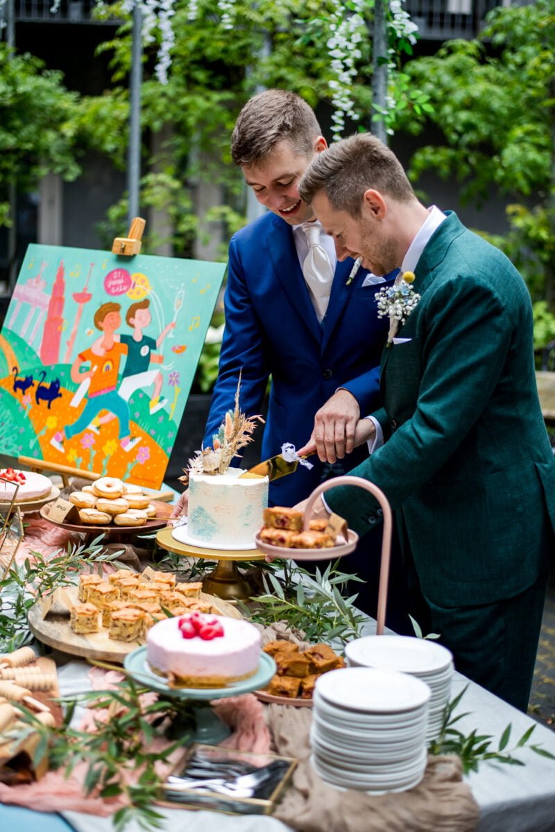 Twee bruidegommen in formele kledij snijden een taart aan tijdens een huwelijksreceptie buiten. De een draagt een blauw pak, de ander een groen pak met een boutonnière. Een kleurrijk schilderij en een assortiment desserts staan op tafel. Borden en bloemdecoraties zijn ook zichtbaar. Weelderig groen vormt het decor.