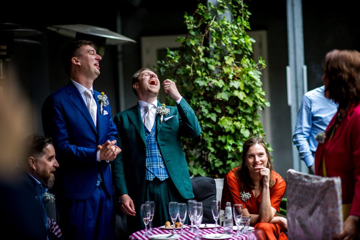 Twee mannen, de een in een blauw pak en de ander in een groen pak en een geruit vest, lachen met hun hoofd naar achteren tijdens een bijeenkomst in de buitenlucht. Een vrouw in een oranje jurk die naast hen zit, glimlacht. Lege champagneglazen staan op een tafeltje bedekt met een rood en wit tafelkleed. Andere mensen zijn gedeeltelijk zichtbaar.
