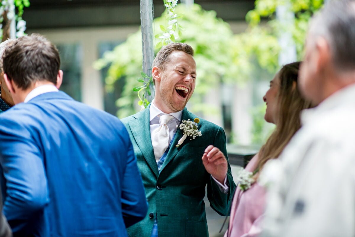 Een man met een groen colbert, wit overhemd en stropdas lacht hartelijk terwijl hij met een vrouw in een roze blazer praat. Ze bevinden zich in een buitenomgeving met groen zichtbaar op de achtergrond. Op de voorgrond staat een man in een blauw pak afgewend. Zowel de lachende man als de vrouw hebben boutonnières.