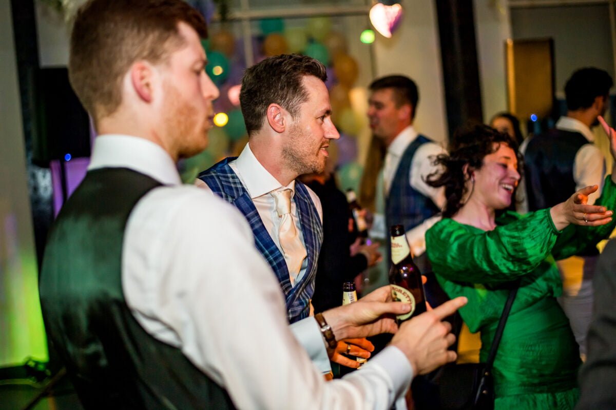Bruidloftfeest in Groningen met vrolijke gasten in nette kleding, bierflesjes, vrouw in groene jurk en kleurrijke ballonnen onder sfeervolle feestverlichting.