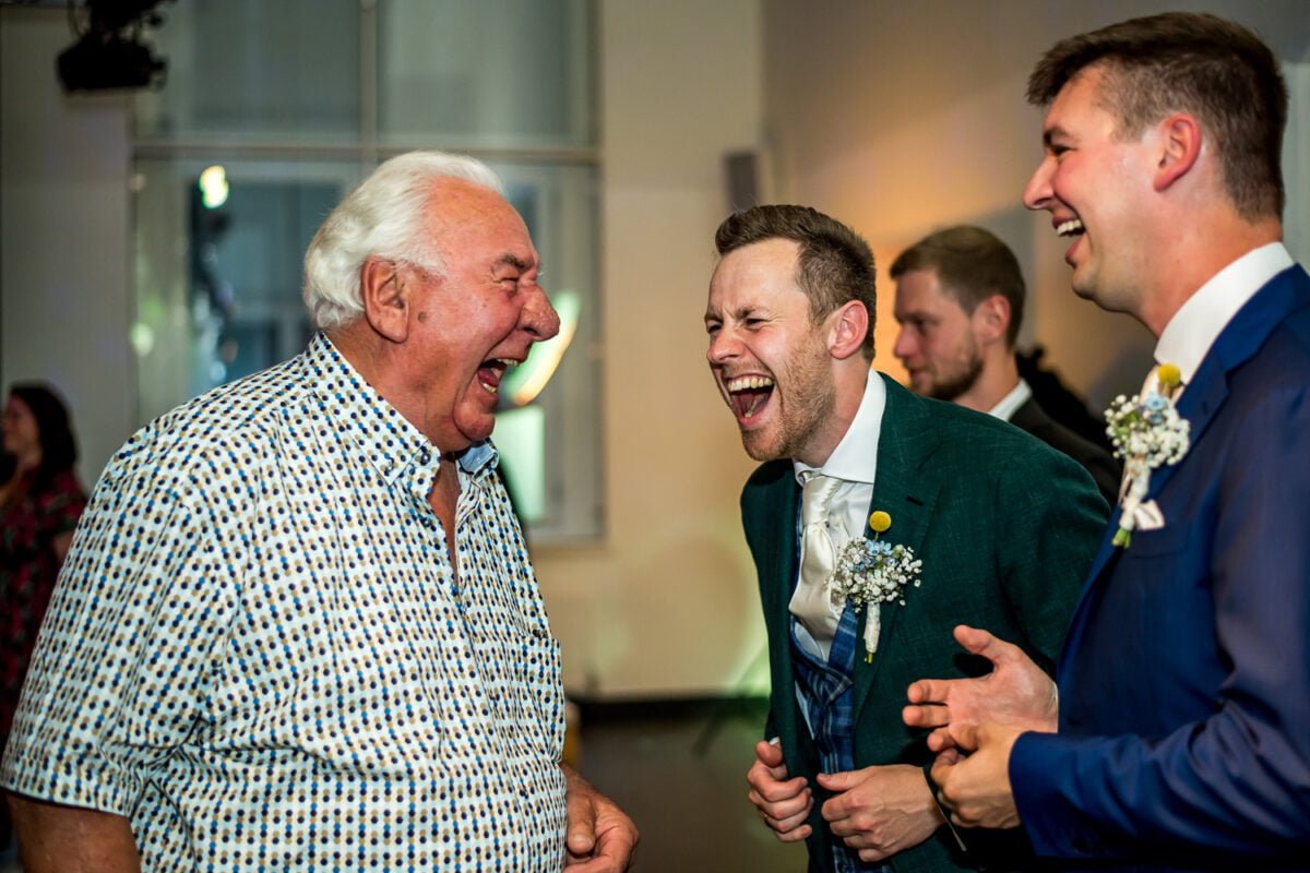 Drie mannen in functionele kleding lachen samen op een bruiloft in Groningen. Twee jonge mannen met corsages en één oudere man, vrolijk samengesteld bij betrouwbare indoor feestverlichting.
