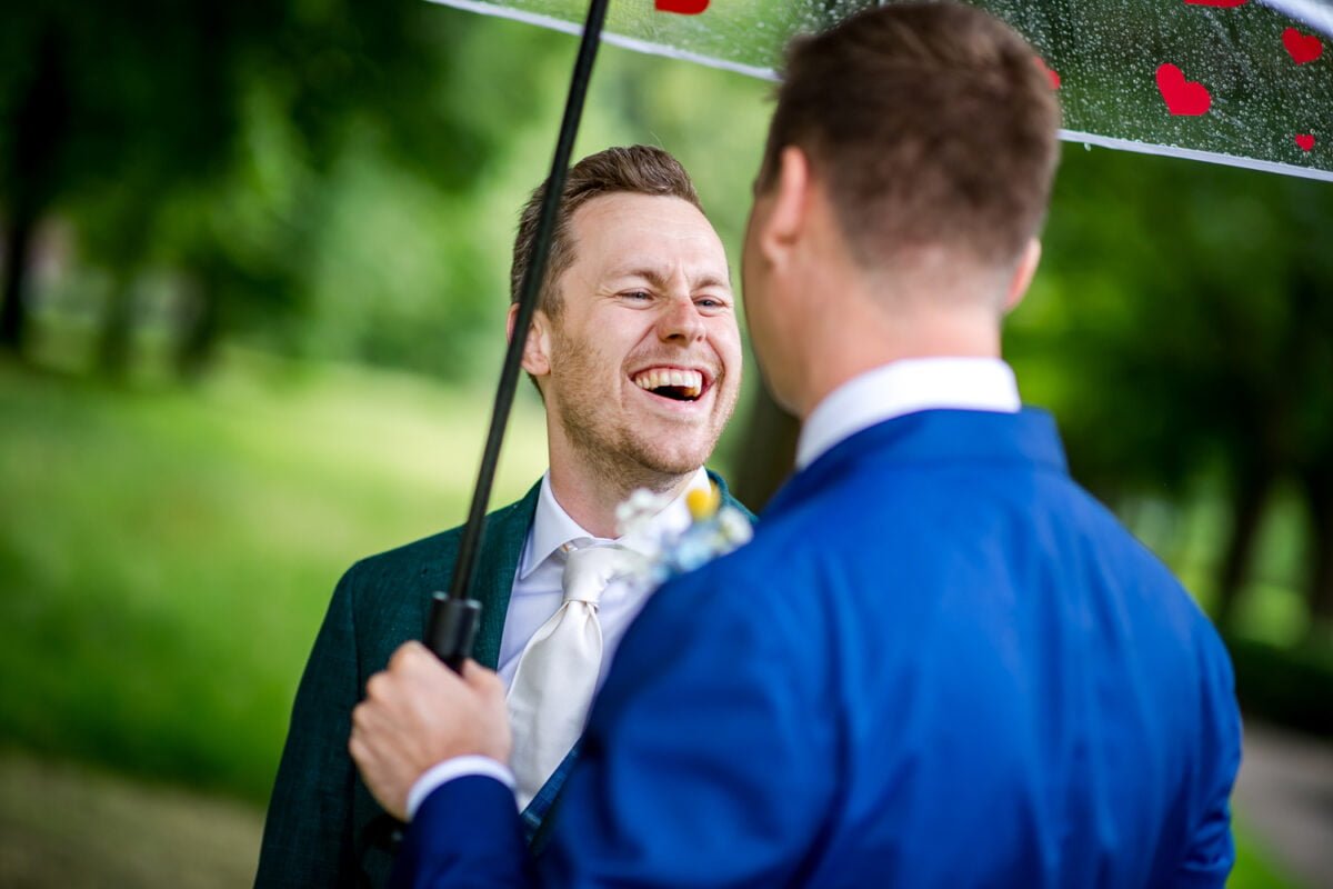 Twee mannen in pak delen een moment buiten onder een paraplu met rode harten. De man links, gekleed in een donkergroen pak en witte stropdas, lacht hartelijk. De man rechts, weggekeerd van de camera, draagt een blauw pak. De setting is een weelderig, groen park.