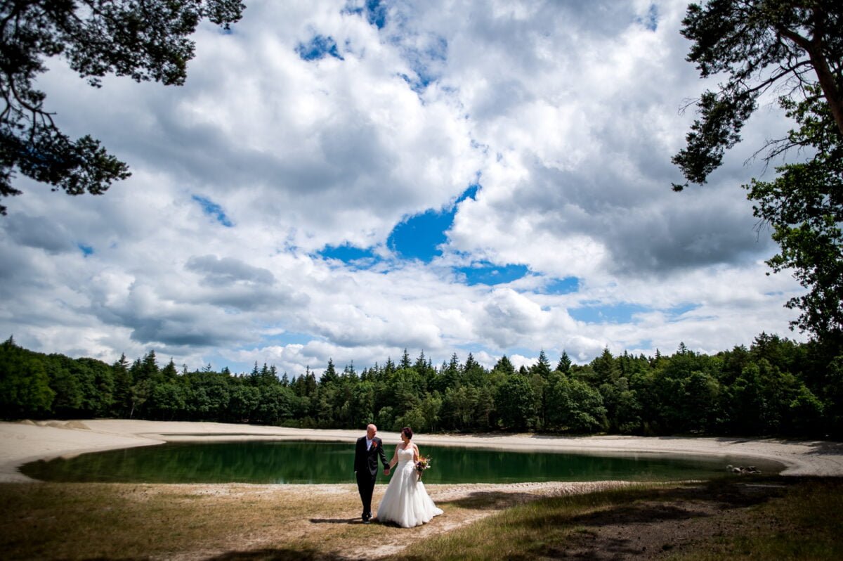 Bruid en bruidstaart wandelen hand in hand bij een rond meer met zandstrand, omringd door groene bomen en een dramatische lucht tijdens hun romantische bruiloft bij de Kruimel.
