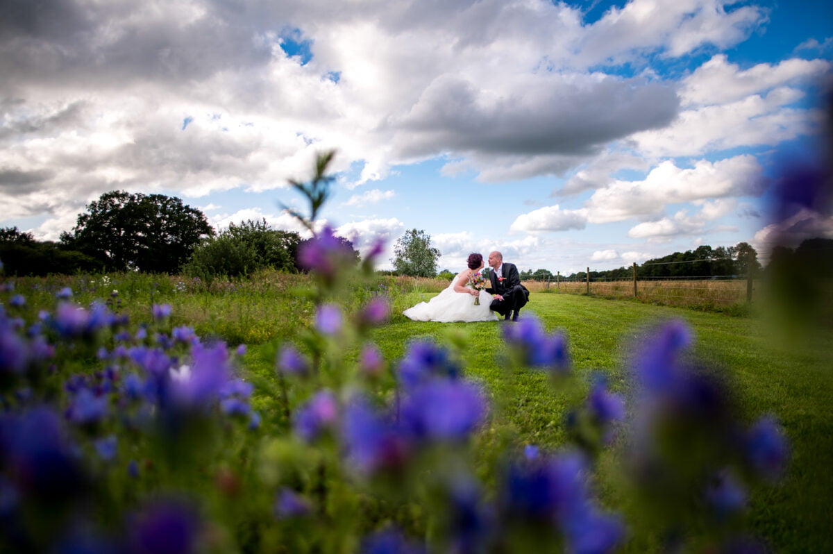 Stellt knie samen op grasveld met paarse bloemen, bomen en hekwerk op de achtergrond, romantische bruiloft bij De Kruimel in landelijk landschap onder deels bewolkte hemel.