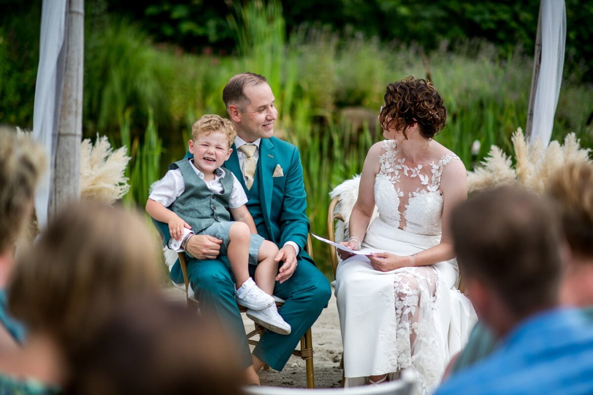 Bruid in witte kanten jurk en bruid in blauwgroen pak lachen samen met jongen op schoot bij buitenbruiloft, omgeven door groen, siergrassen en gasten bij de Kruimel.