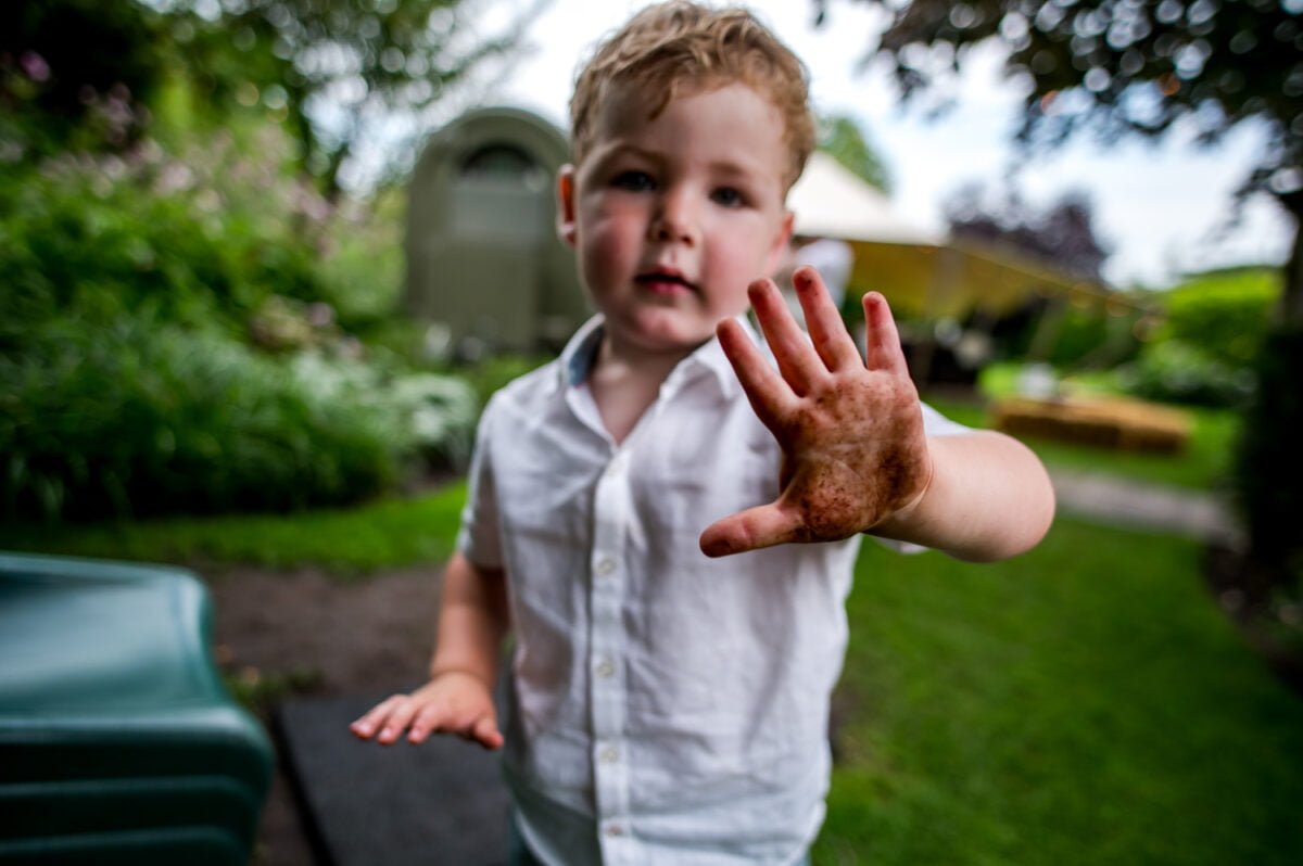 Kind met krullend blond haar toont modderige hand buiten op gras bij de Kruimel, naast door bomen en tuin in een parkachtige omgeving. Buiten spelen, vieze handen, natuur.