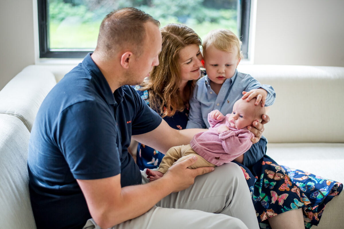 Jong gezin met peuter en pasgeboren dochtertje zittend op een witte bank, samen thuis met natuurlijk licht en groen buiten het raam. Liefdevolle ouders verwelkomen hun nieuwe broertje of zusje.