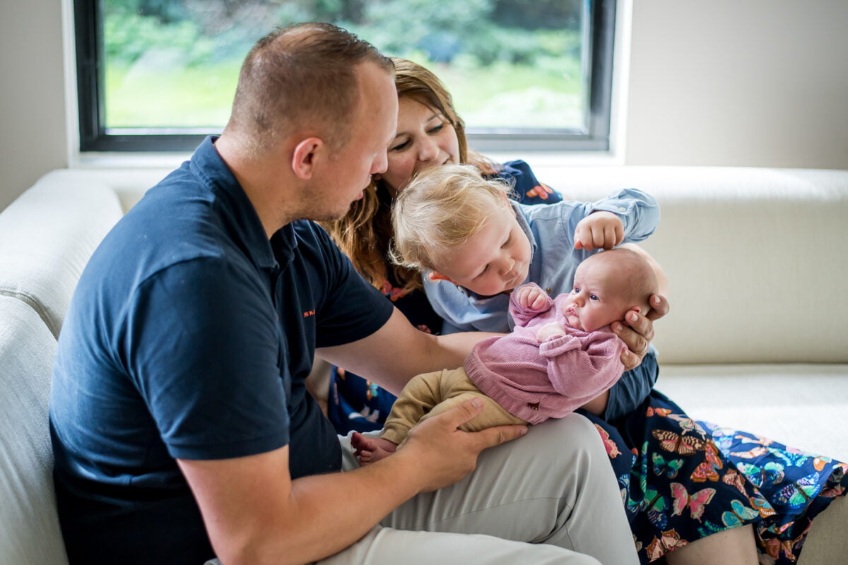 Gezin met pasgeboren baby en peuter op witte bank, omringd door warm daglicht uit groot raam. Liefdevolle interactie, samen genieten van gezinsgeluk en nieuw leven.