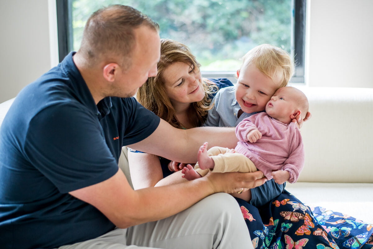 Gezin met twee jonge kinderen op witte bank, moeder glimlacht terwijl peuter de pasgeboren baby in roze knuffelt, vader ondersteunt het hoofdje; raam en groen buiten zichtbaar.