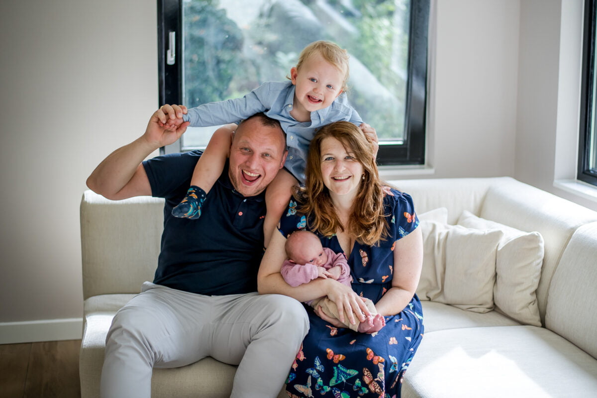 Een gelukkig gezin van vier op een lichte bank, met ouders die hun kinderen vasthouden, zonlicht dat door de ramen stroomt, lichte, gezellige woonkamer.