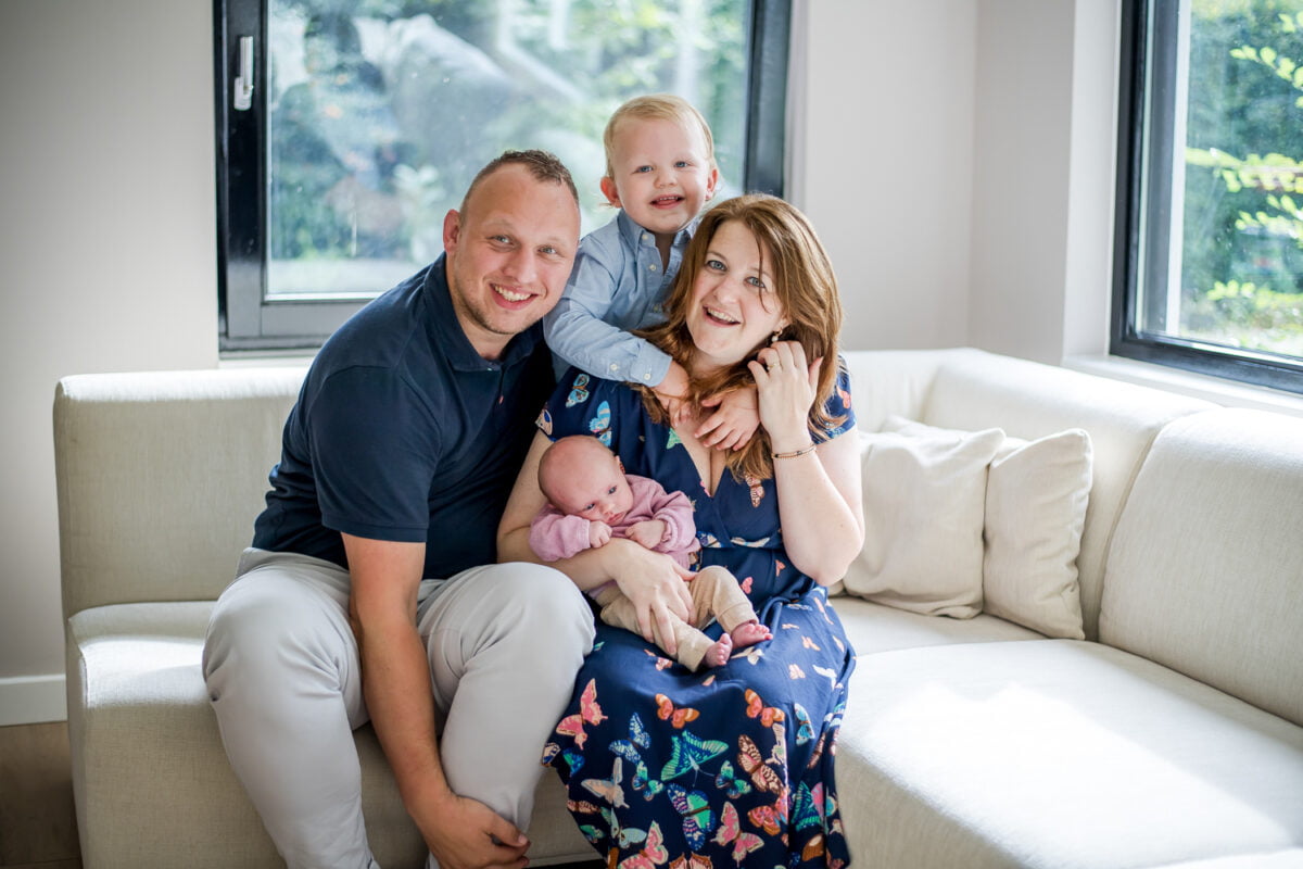 Gezin van vier op crèmekleurige bank in lichte woonkamer met grote ramen; moeder houdt pasgeboren baby vast, ouder kind knuffelt haar, vader erbij, allemaal lastig.