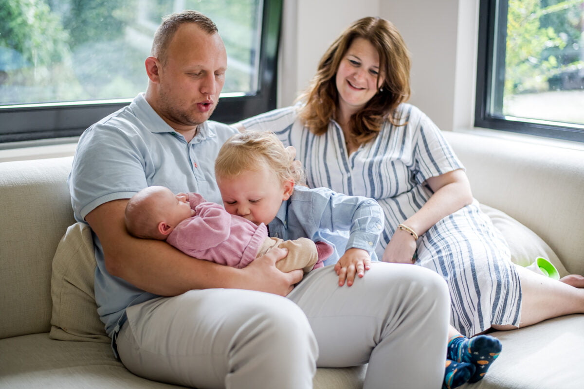 Een gelukkig gezin thuis: vader met pasgeboren baby in roze, peuter die broertje of zusje kust, moeder die lacht op de bank. Zonlicht door de grote ramen zorgt voor een warm, vrolijk familiemoment in de woonkamer.