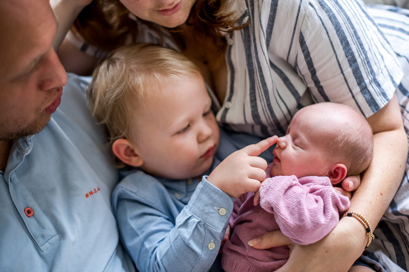 Twee jonge kinderen, inclusief een pasgeboren baby in roze kleding, worden liefdevol omarmd door volwassenen in lichte kleding; intiem familieportret met warme sfeer.