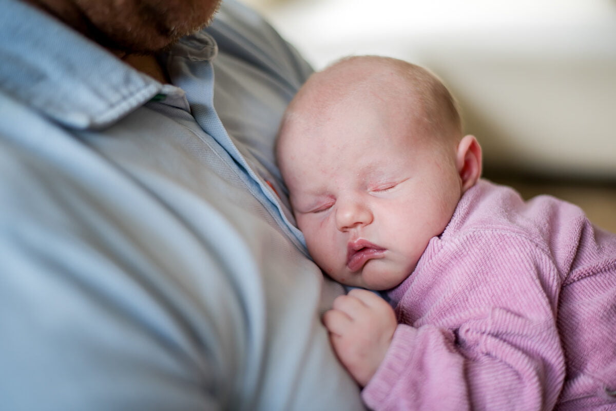 Veroorzaakt baby in roze outfit roest op borst van volwassene met lichtblauw overhemd, handje grijpt shirt, ontspannen gezicht en gesloten ogen, zachte en kalme sfeer.