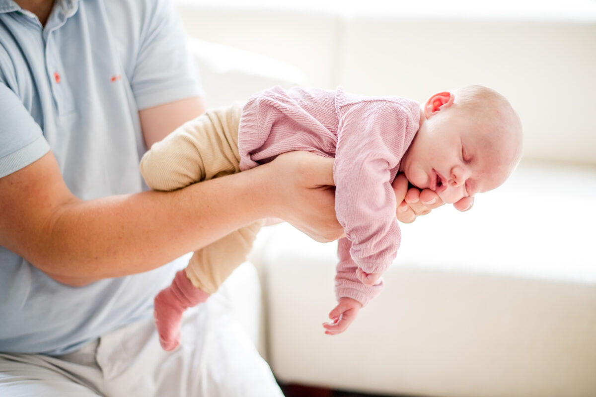 Pasgeboren baby in een roze topje en beige broekje slaapt op de buik, zachtjes gekoesterd door zorgzame handen, gekleed in een lichtblauw shirt, rustige binnenomgeving, pasgeborene verzorging en hechting.