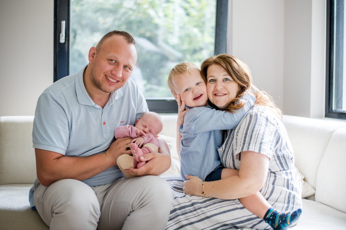 Gezin van vier op witte bank in zonnige woonkamer met grote ramen. Vader houdt van slapende baby vast, moeder knuffelt zoon. Gezinsgeluk en natuurlijk licht, moderne woning, binnenkijken.