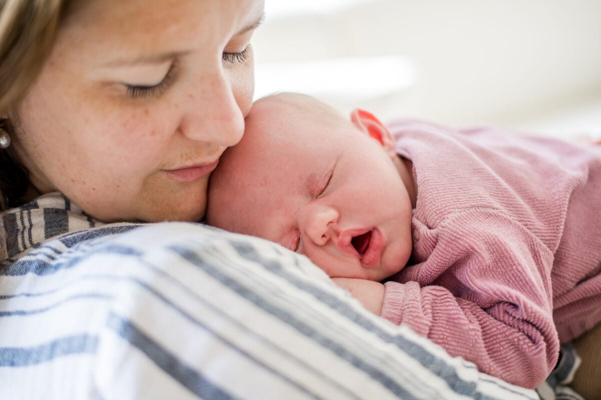 Moeder met lichte huid en sproeten knuffelt pasgeboren baby in roze outfit. Baby slaapt rustig op schouder. Intieme, zacht moeder-kind moment, newborn fotografie.