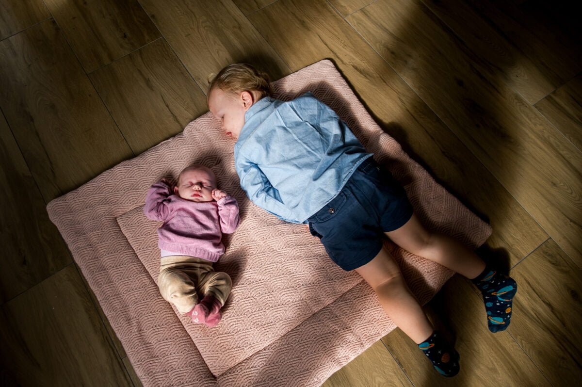 Baby en peuter samen op roze speelmat op houten vloer; pasgeboren baby slaapt in roze kleding, peuter in blauwe outfit kijkt weg – familie en kindertijd concept.