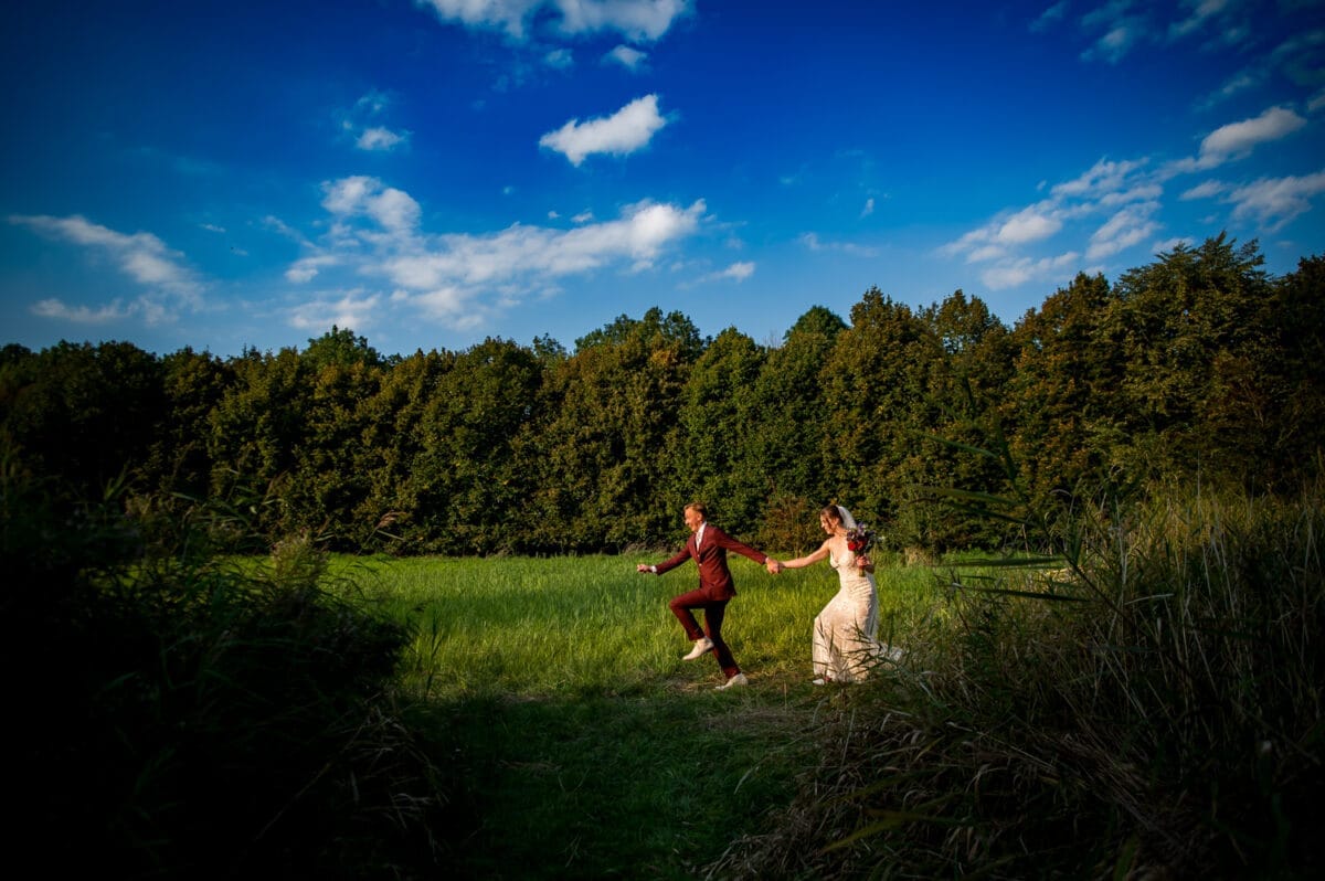 Bruidspaar hand in hand rennend door groen veld bij Rietland, bruiloft onder blauwe lucht met wolken, hoge bomen op de achtergrond, gras en liefdevolle sfeer.