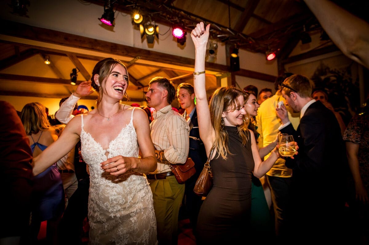 Bruiloft dansende bruid in witte kanten jurk omringd door feestende gasten, kleurrijke lichten en houten balken in sfeervolle Rietland trouwlocatie.