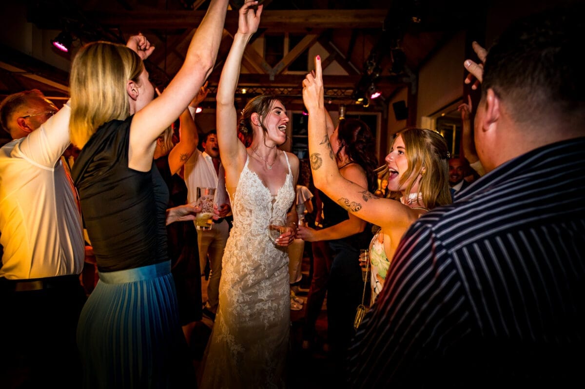 Bruiloft bij Rietland: bruid in witte kanten jurk danst met gasten, feestelijke sfeer, vrolijke mensen zingen en vieren onder warme verlichting.