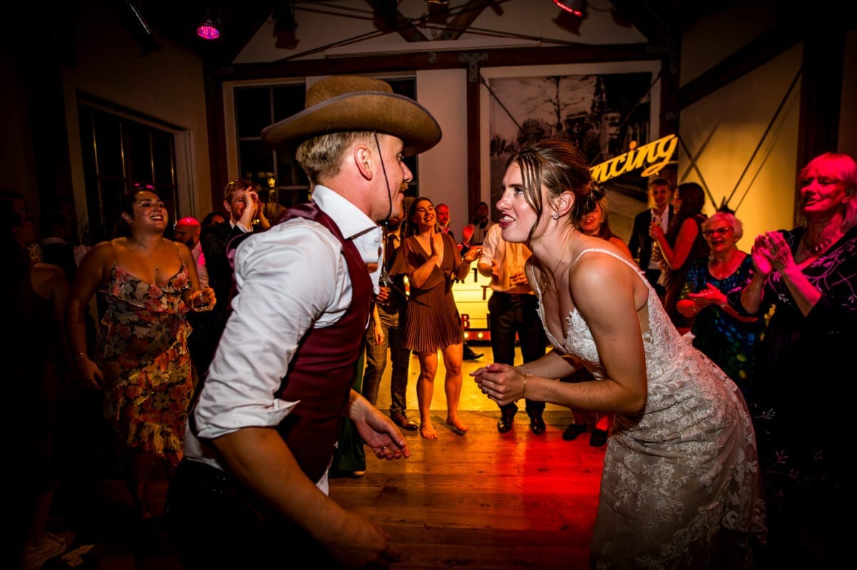 Bruiloft Rietland: bruid in witte kanten jurk en man met cowboyhoed dansen energiek op houten vloer, omgeven door lachende gasten in kleurrijke feestkleding.