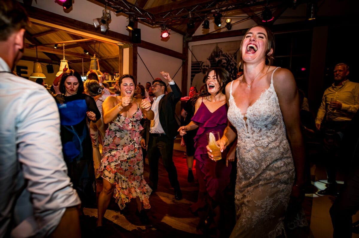 Dansende gasten op een vrolijke indoor bruiloft bij Rietland, vrouw in witte kanten trouwjurk vooraan, feestelijke verlichting, sfeer van geluk en energie tijdens huwelijksviering.