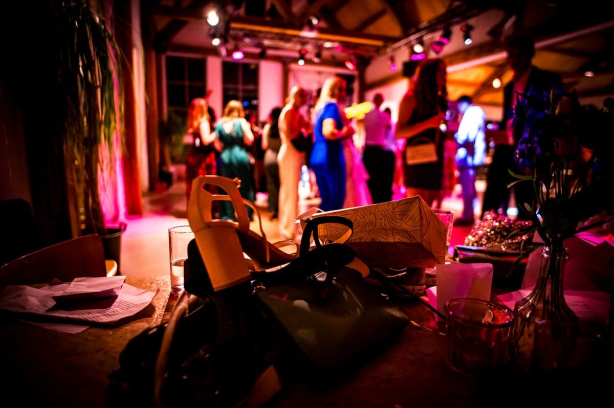 Bruiloftsfeest bij Rietland: feestvierders dansen onder roze en paarse lichten, hoge plafonds; op de voorgrond een volle tafel met tassen, glazen en papieren in een sfeervolle feestzaal.