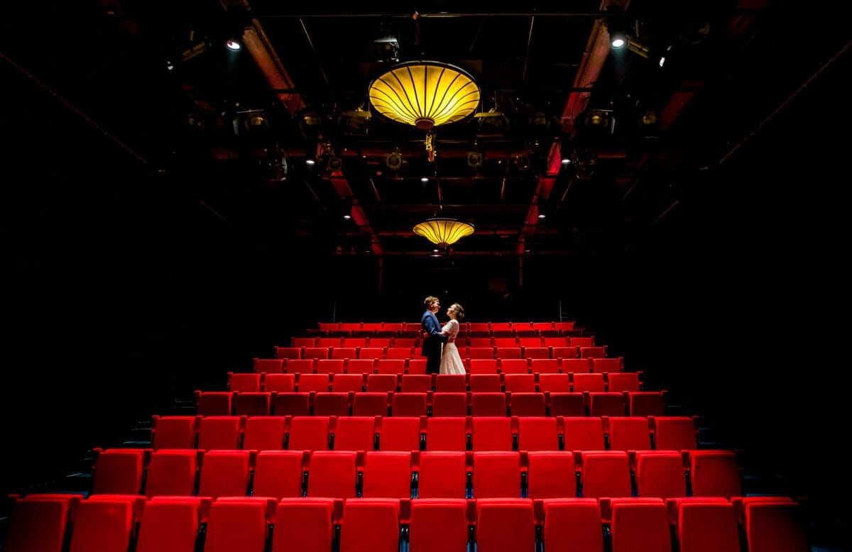 Stel in galakleding omarmt elkaar op de trappen in een lege theaterzaal met rode stoelen en warme gele verlichting, romantische sfeer, avondje uit, theaterinterieur.