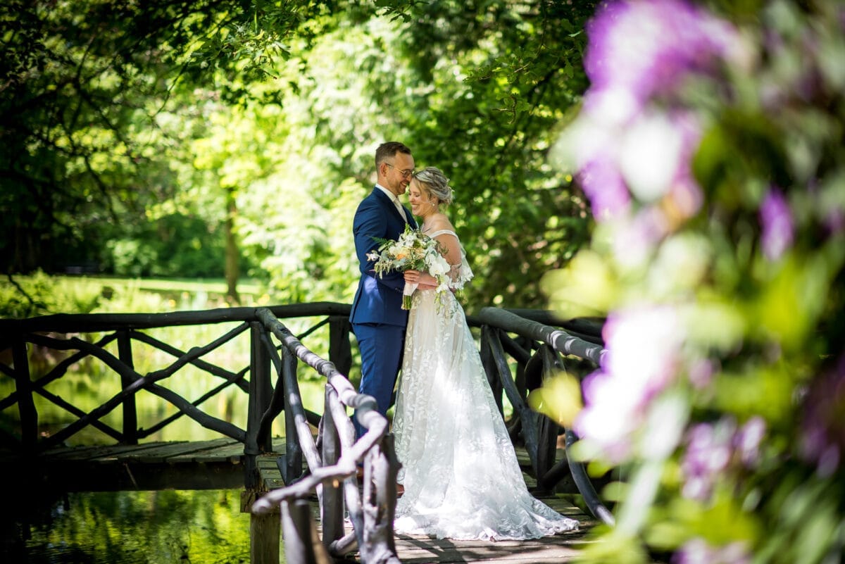 Bruiloftspaar op houten brug in groen park, bruid met boeket in witte jurk, bruid in blauw pak. Zonlicht door bomen, paarse bloemen scherp op de voorgrond.